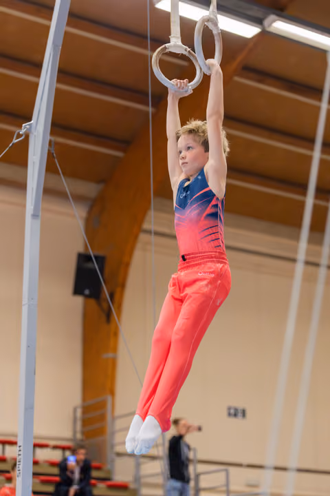 Young male gymnast hangs from still rings with focused expression, wearing red and blue leotard in a sports hall.