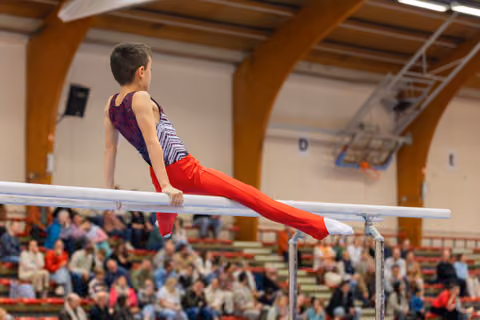 Young male gymnast holds a straddle position on parallel bars, focused and controlled, before a packed gymnasium crowd.