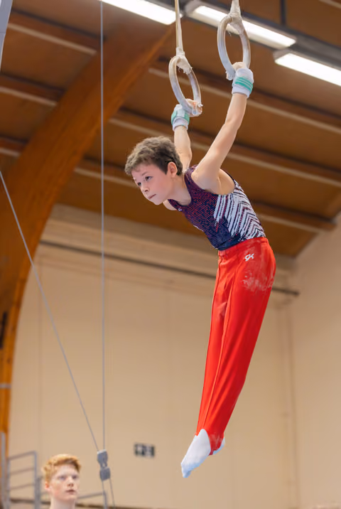 Young male gymnast hangs focused on still rings, red pants and wrist guards, in a wooden-ceiling gymnasium.
