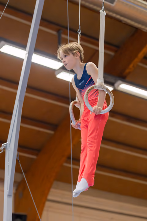 Young gymnast hangs with focused concentration on still rings in a wooden-beamed gymnasium, legs crossed mid-routine.
