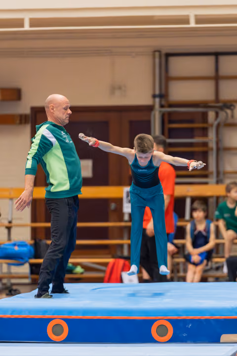 Bald coach in green jacket steadies a focused young gymnast mid-air above a blue trampoline mat indoors.