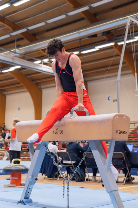 Young male gymnast in red and black leotard performs on pommel horse, concentrating intensely during competition.