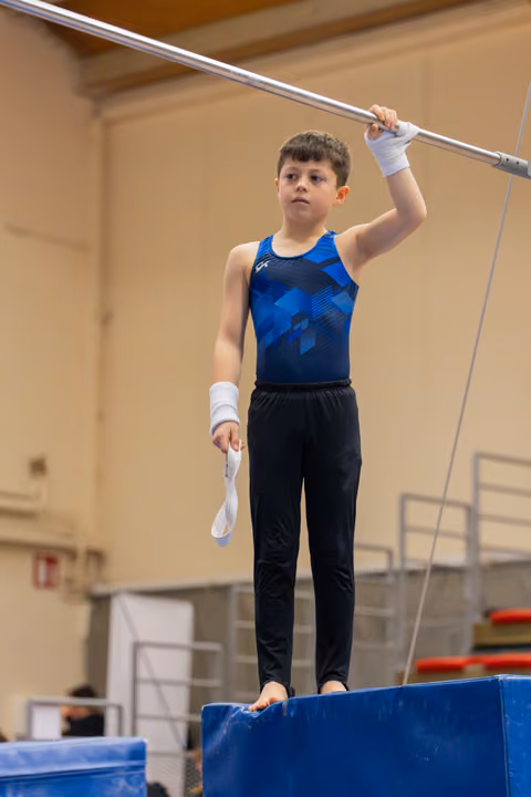 Young boy gymnast stands barefoot on a block, gripping the high bar with focused concentration, wearing blue leo and wrist wraps.