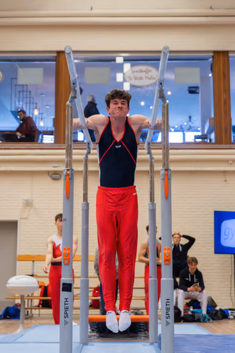 Young male gymnast grips parallel bars mid-routine, face showing intense concentration, wearing red and black leotard indoors.