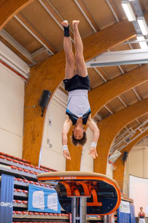 Young male gymnast performs a handstand above a Spieth vault, body fully extended, in an indoor gymnastics arena.