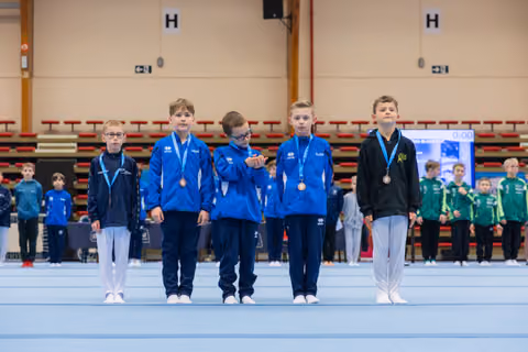 Five young boys wearing bronze medals stand proudly on the competition floor, dressed in blue tracksuits, inside a gymnasium.