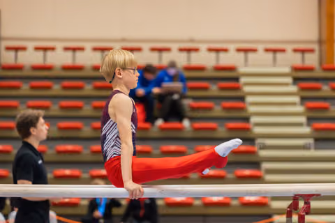 Blond boy with glasses performs a straddle hold on parallel bars, focused and composed, in a gymnastics hall.
