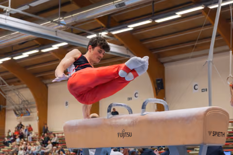 Young male gymnast swings his legs powerfully over a Spieth pommel horse, focused expression, indoor gymnastics hall.