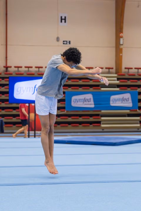 Young male gymnast leaps mid-air with arms tucked forward, focused expression, on blue floor at Gymfed competition.