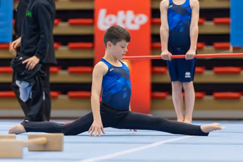 Focused young male gymnast performs a side split on the floor mat, eyes downcast, during a gymnastics event.