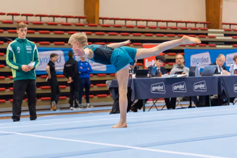 Young gymnast performs a precise arabesque on the floor, watched by a coach in green and judges at the Gymfed table.