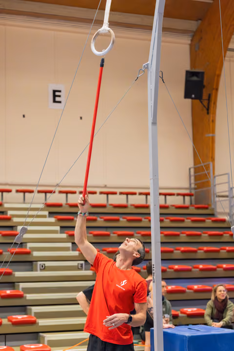 Coach in red shirt uses a long red pole to position gymnastics rings, gazing upward with focus in an indoor arena.
