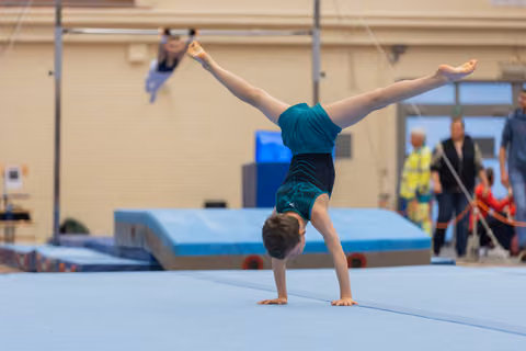 Young gymnast performs a perfect handstand with legs in a full split on a blue gymnastics mat during a competition.