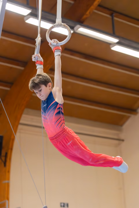 Young boy in red leotard swings on gymnastic rings with intense focus, body arched gracefully in a gym.