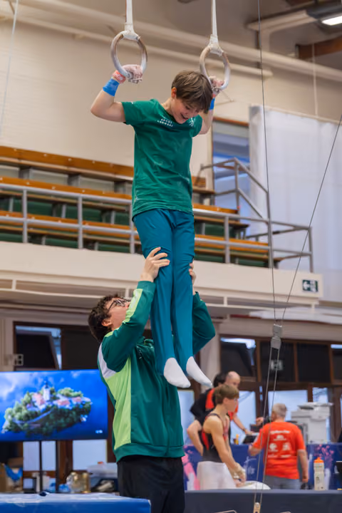 Smiling boy in green hangs from gymnastics rings while a coach steadies his legs in a busy sports hall.