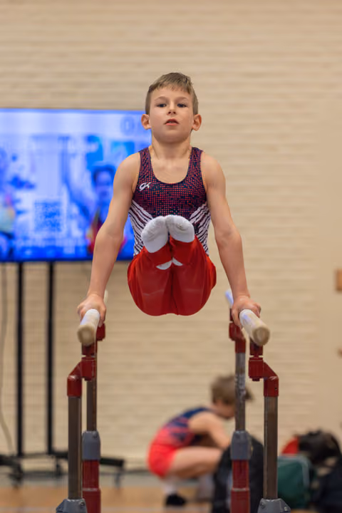 Young male gymnast holds a straddle position on parallel bars, focused expression, wearing red shorts and GK leotard.