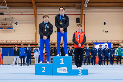 Three young gymnasts stand on a Gymfed podium; the gold medalist stands tall in the center, flanked by silver and bronze winners.