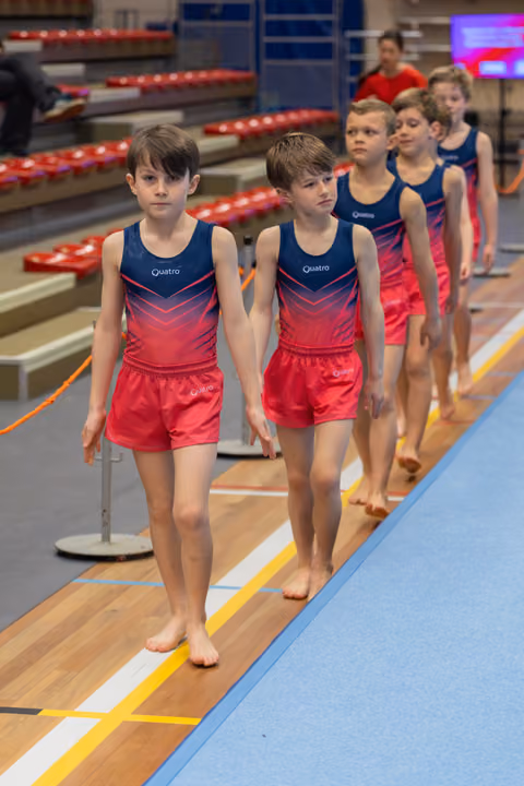A line of young male gymnasts in matching Quatro uniforms walk barefoot across the floor, focused and composed.