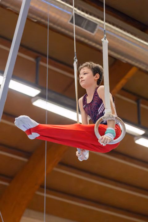 Young boy performs on gymnastics rings with focused expression, legs extended, wearing red pants in a wood-beamed gym.