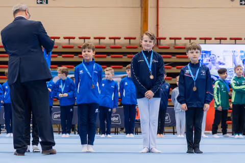 Three young gymnasts stand proudly on the podium wearing bronze medals, while an official presents awards at a gymnastics meet.