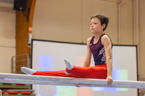 Focused young male gymnast performs a straddle hold on parallel bars, gazing ahead with calm determination in a sports hall.
