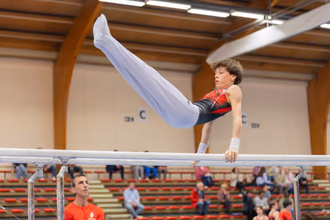 Young male gymnast performs a straddle hold on parallel bars, focused and controlled, as a coach watches below.