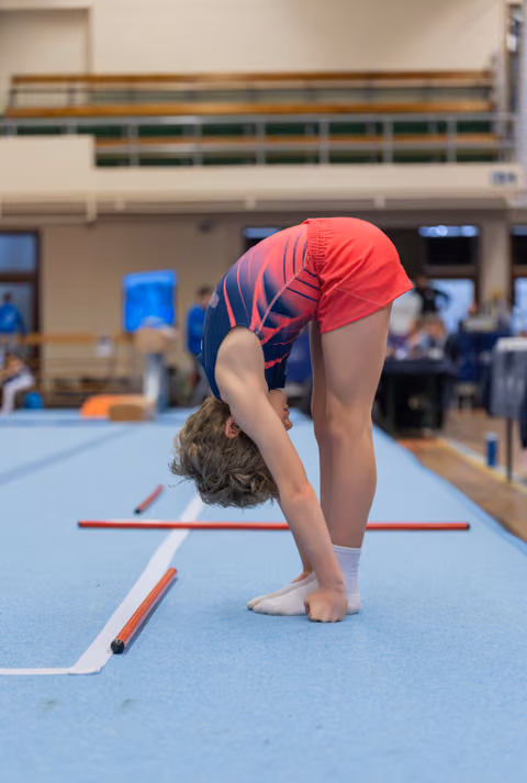 Young gymnast bending forward in a deep toe touch on the blue floor exercise mat during an indoor gymnastics meet.