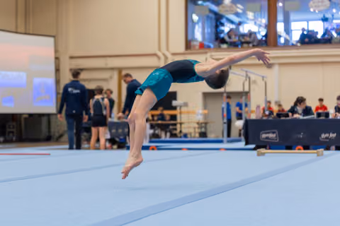 Young gymnast in teal leotard executes a powerful backflip on the floor exercise mat during an indoor competition.
