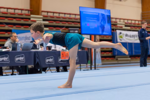 Young boy balances on one leg in an arabesque pose on the floor exercise mat, focused and controlled at a gymnastics meet.