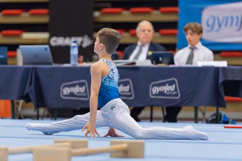 Young male gymnast performs a split on the floor, focused and poised, while judges observe from a table behind him.