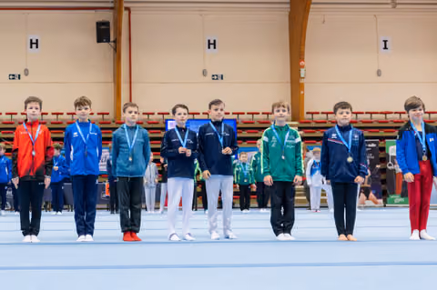 Eight young male gymnasts stand proudly on the competition floor, wearing medals around their necks at an indoor gymnastics event.