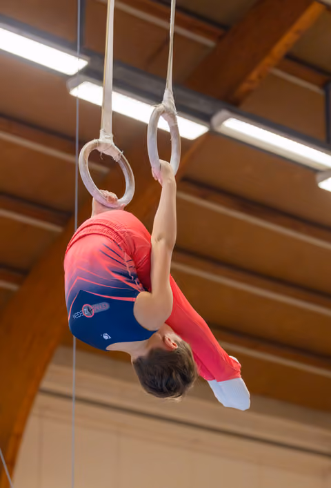 Young male gymnast hangs inverted on still rings, body curled, focused effort visible in indoor gymnasium.