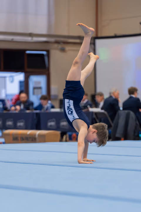 Young boy performs a precise handstand on the floor exercise mat, legs extended upward, during an indoor gymnastics competition.