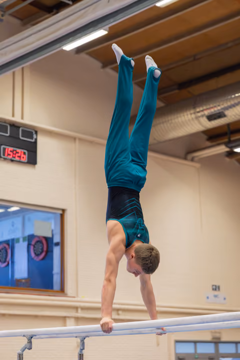 Young male gymnast holds a perfect handstand on parallel bars, legs split, in a gym with a digital clock visible.