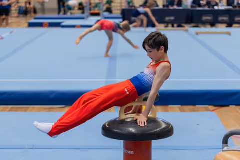 Young male gymnast holds a tuck position on a mushroom pommel, face intent and focused, in a busy competition gym.