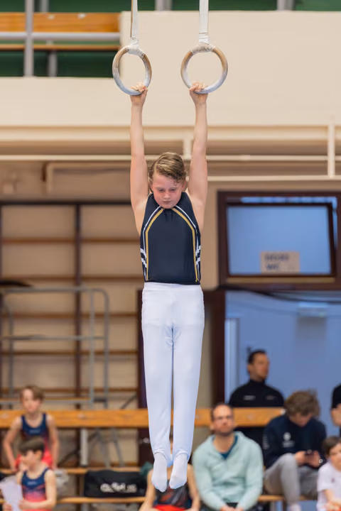 Young male gymnast hangs from still rings with focused concentration, arms extended overhead, in a gymnasium setting.