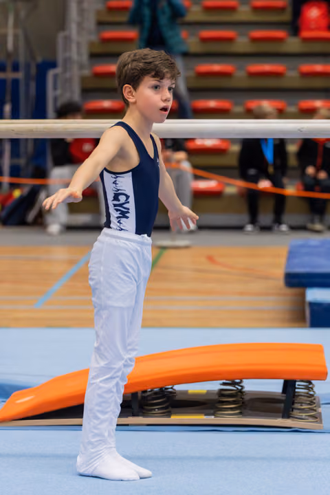 Young boy in navy leotard stands alert by springboard, arms slightly raised, focused expression in a gymnastics arena.