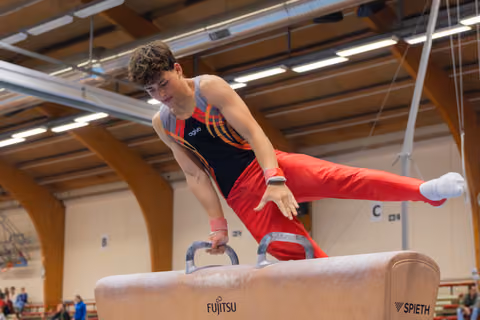 Young male gymnast performs on pommel horse, leaning forward with intense focus, legs extended mid-routine in indoor gym.