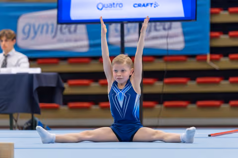 Young gymnast performs splits with arms raised high, focused expression, during a gymnastics meet on a blue floor.