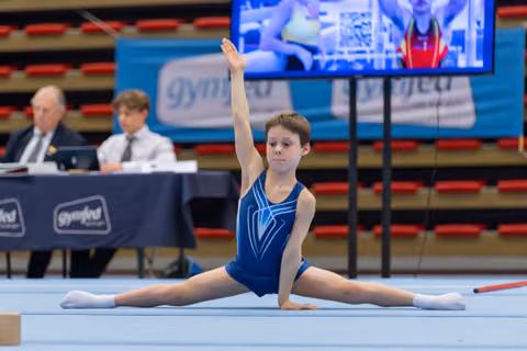 Young gymnast performs a split on the floor exercise mat, arm raised, focused expression, judges seated in background.