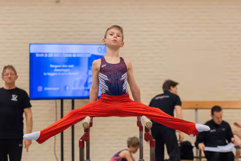 Young boy performs a straddle hold on parallel bars, legs split wide, with a focused, confident expression.