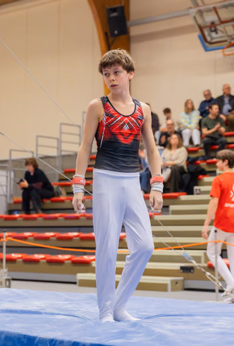 Young male gymnast in red leotard stands composed on blue mat, wrists taped, spectators watching in background.