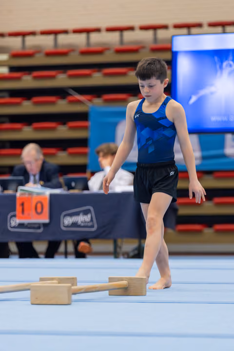 Focused young male gymnast walks barefoot on blue floor, studying wooden apparatus blocks, judges visible behind him.