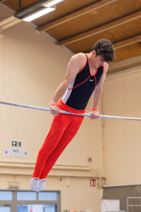Young male gymnast grips the high bar, leaning forward with focused concentration in a gymnasium setting.
