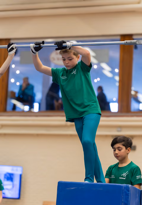 Focused boy in green grips horizontal bar standing on blue block, younger teammate watches attentively below.