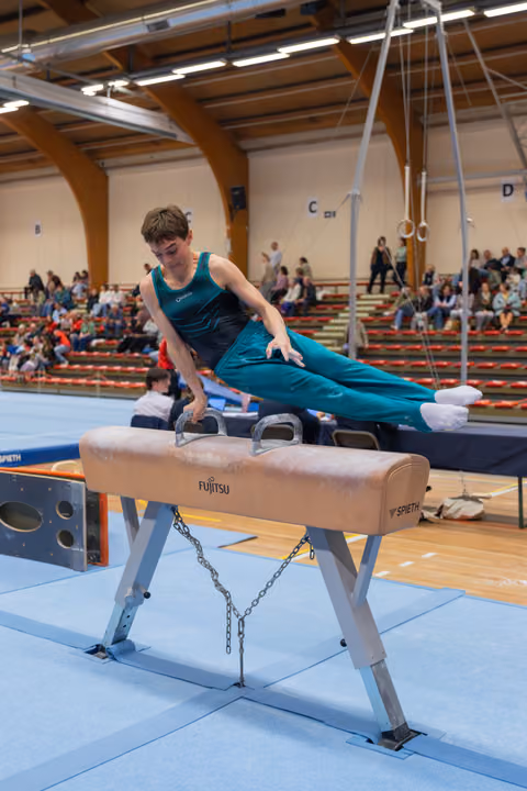 Focused teenage gymnast in teal leotard performs pommel horse routine, body extended horizontally, in packed sports hall.