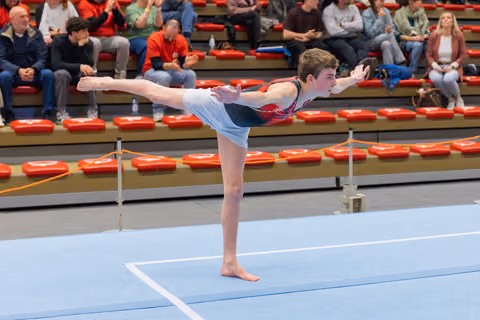Teen male gymnast holds a precise arabesque on the floor, arms forward, focused expression, crowd watching in bleachers.