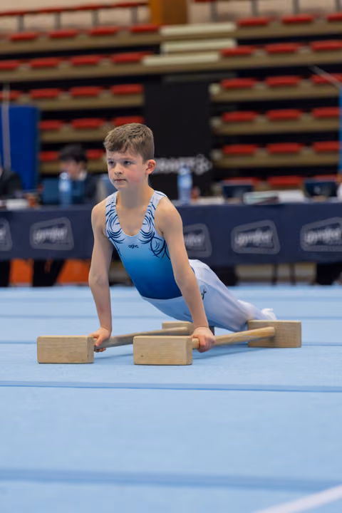 Young boy in blue leotard performs floor exercise on parallettes, focused expression, at indoor gymnastics event.
