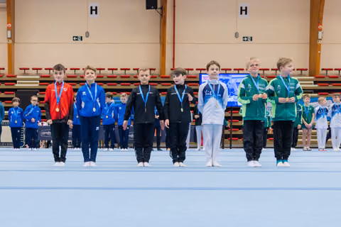 Young boys lined up on the gymnastics floor wearing medals, displaying mixed emotions from proud smiles to neutral expressions.