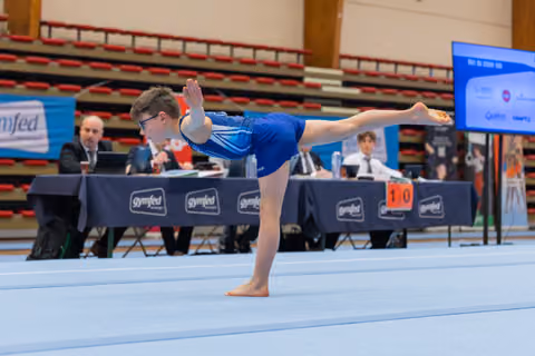 Young boy with glasses performs a focused arabesque on the floor, judges seated at gymfed table behind him.